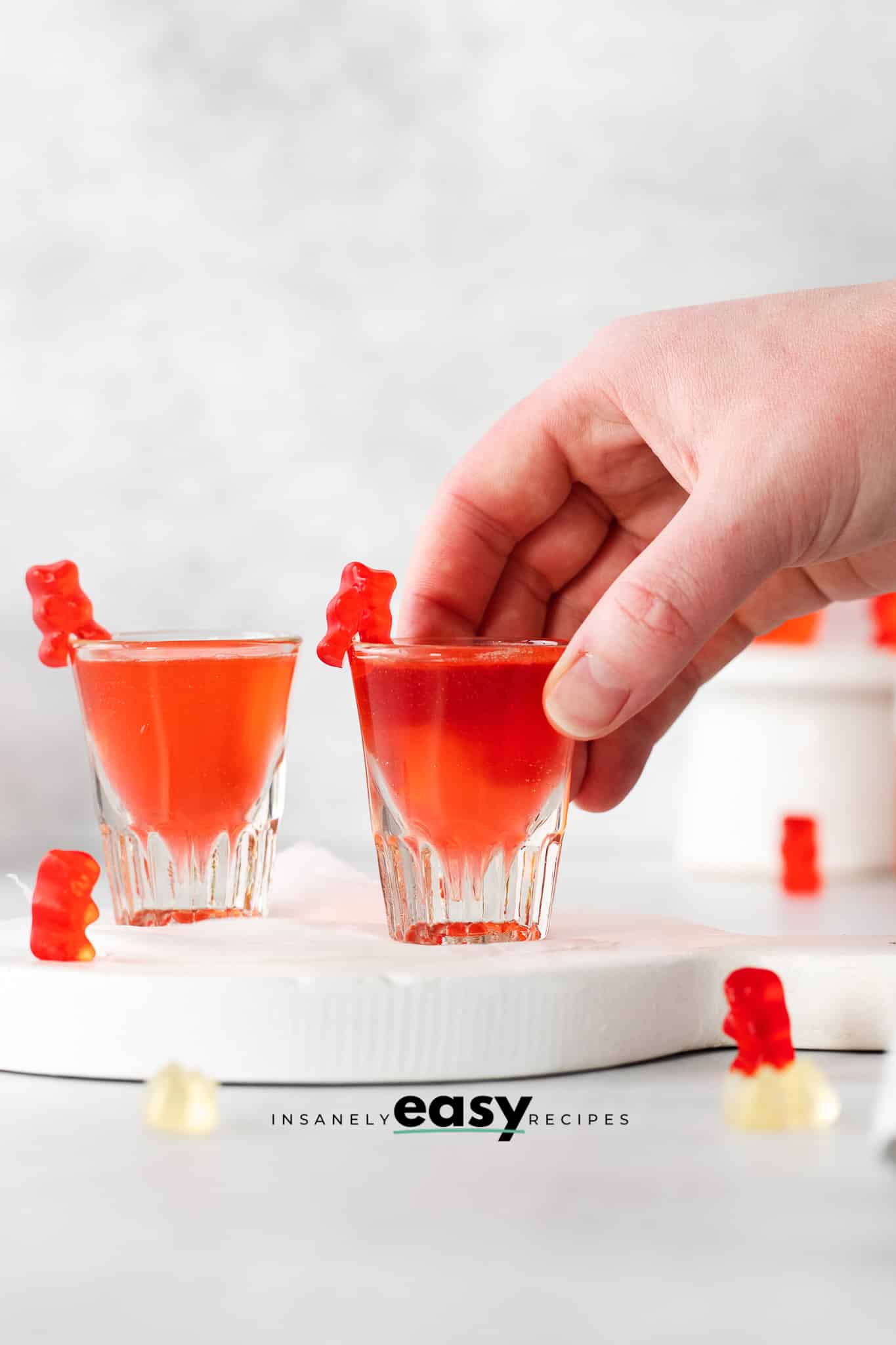 clear shot glasses filled with red liquid and a red gummy beaer on top, some gummy bears scattered on the table.