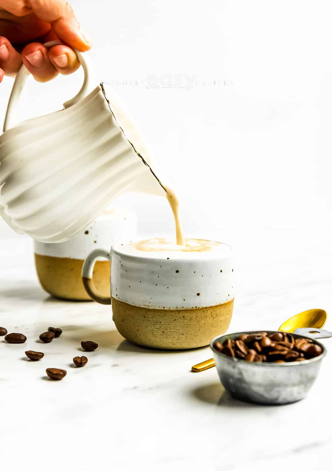 Photo of a hand pouring oat milk from a small white pitcher into a mug filled with coffee. There is a measuring cup filled with coffee beans and a gold spoon in the foreground and a mug in the background. 