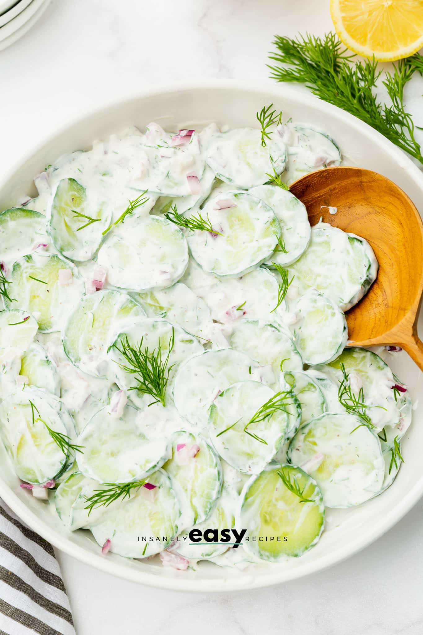 Sliced cucumbers dressed in sour cream for polish cucumber salad. a large wooden spoon is serving the salad, and fresh dill sprigs are sprinkled on.