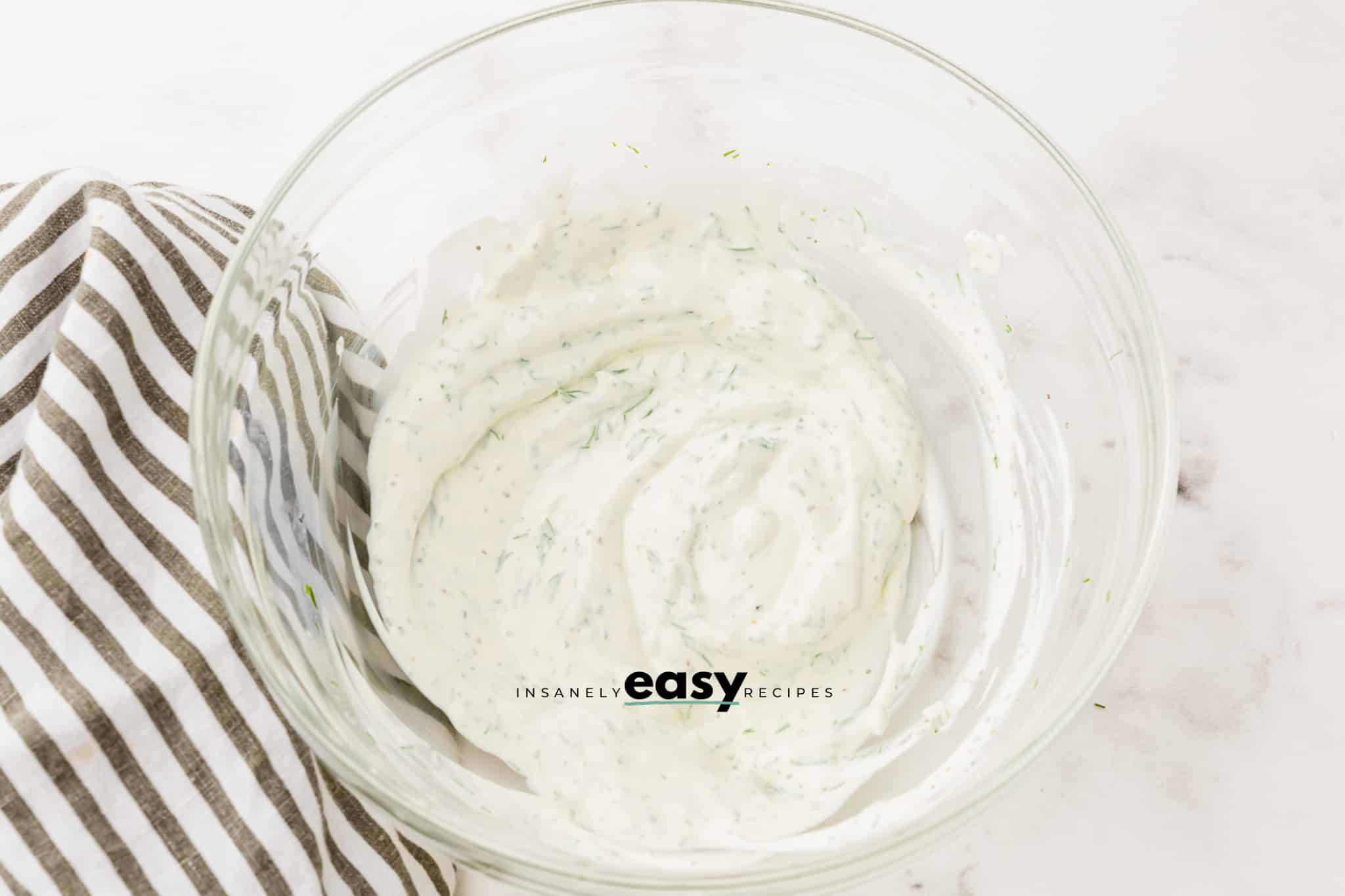 A glass mixing bowl with sour cream and seasonings for cucumber salad.
