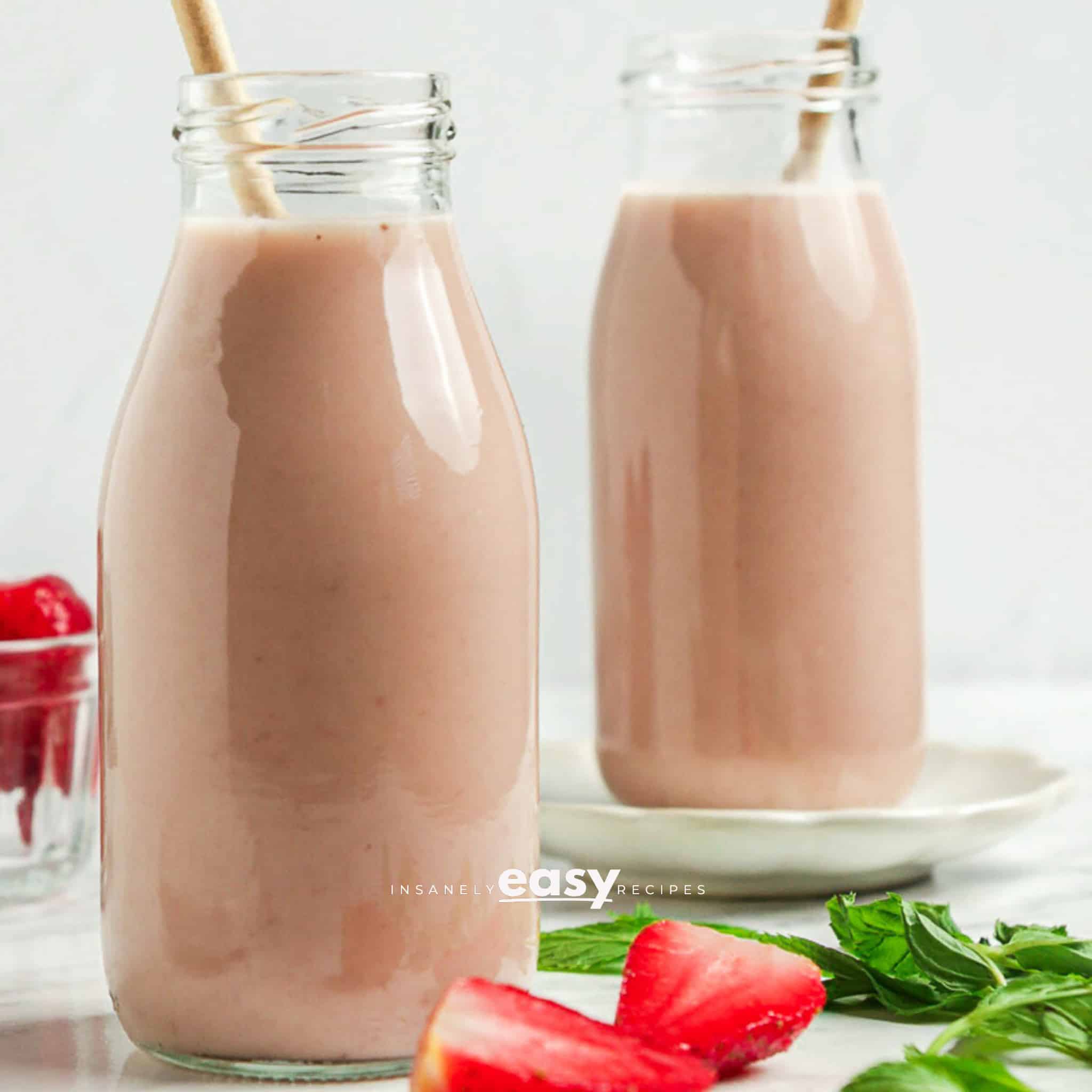 Photo of two tall glass jars filled with Strawberry Almond Milk. There are strawberries by the jars and straws in the jars.