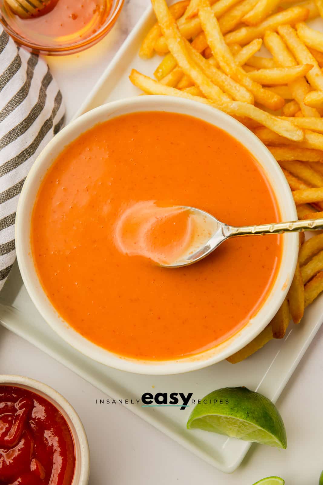 orange sauce in white bowl surrounded by fries and silver spoon in sauce