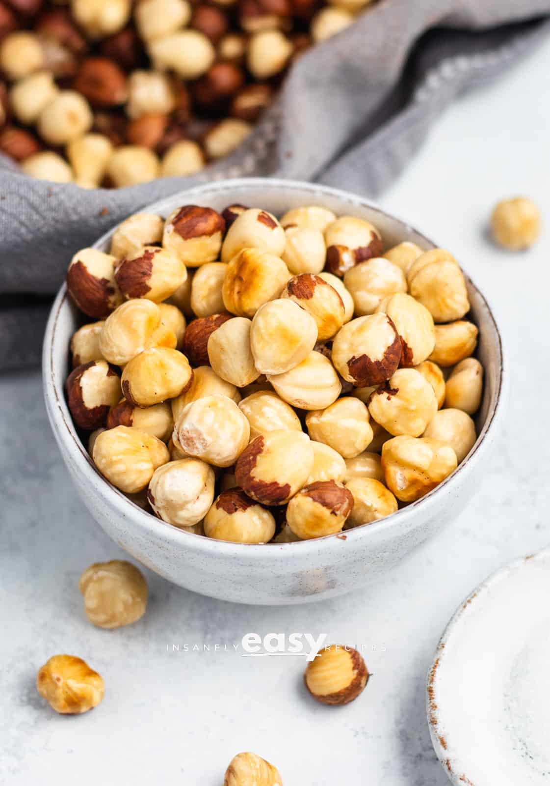Photo of roasted and peeled hazelnuts in a small bowl, with a towel filled with hazelnuts in the background