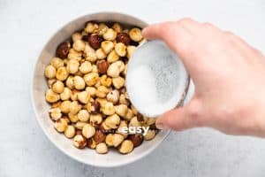 Top view photo of a hand sprinkling sauce into a bowl of peeled and roasted hazelnuts.