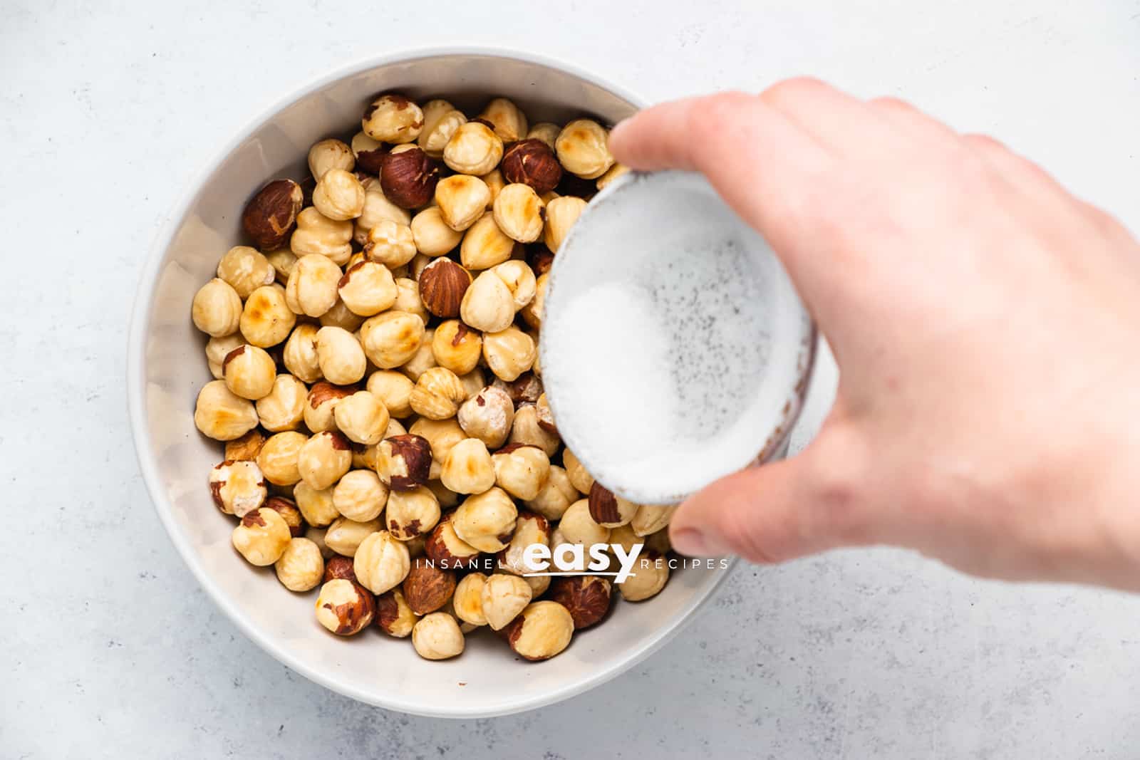 Top view photo of a hand sprinkling sauce into a bowl of peeled and roasted hazelnuts.