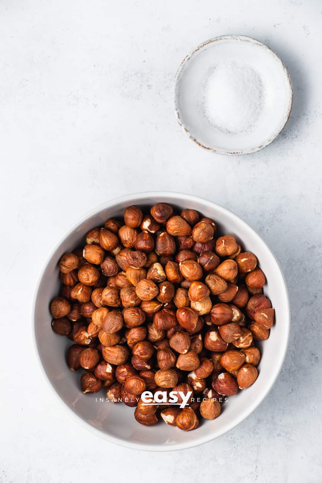 Top view photo of unroasted hazelnuts and salt in separate bowls.