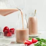 Photo of a hand pouring Strawberry Almond Milk in a tall glass bottle. There is another bottle of Strawberry Almond Milk in the background with a straw and strawberries around the jars.