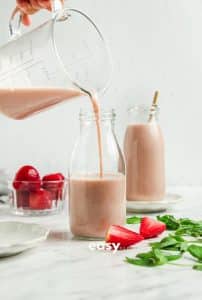 Photo of a hand pouring Strawberry Almond Milk in a tall glass bottle. There is another bottle of Strawberry Almond Milk in the background with a straw and strawberries around the jars.