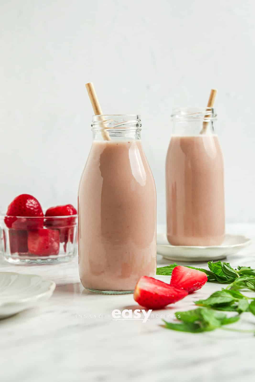 Photo of two tall glass jars filled with Strawberry Almond Milk. There are strawberries by the jars and straws in the jars. 