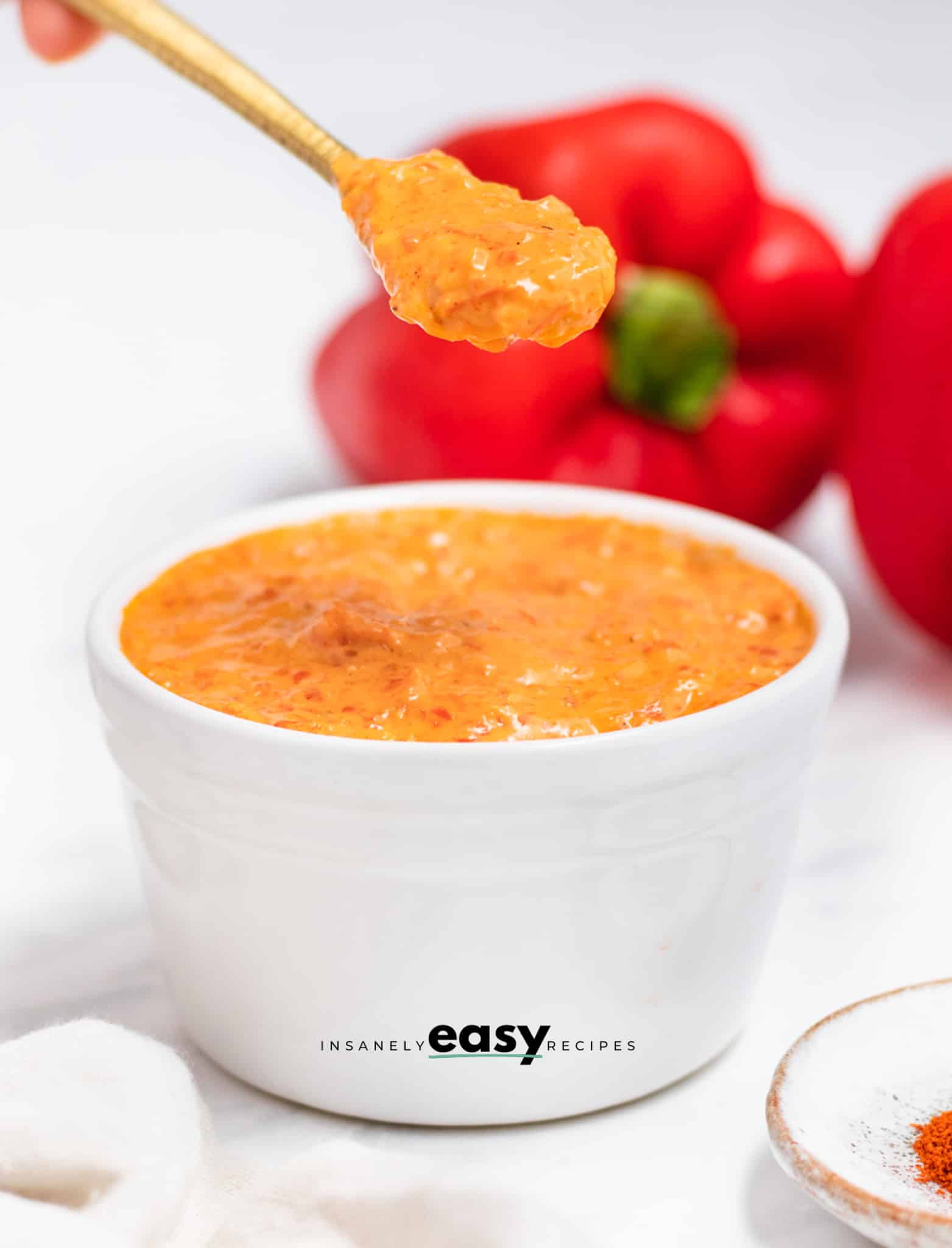 a bowl of red pepper crema. a spoon is lifting some up. In the background are fresh red bell peppers. 