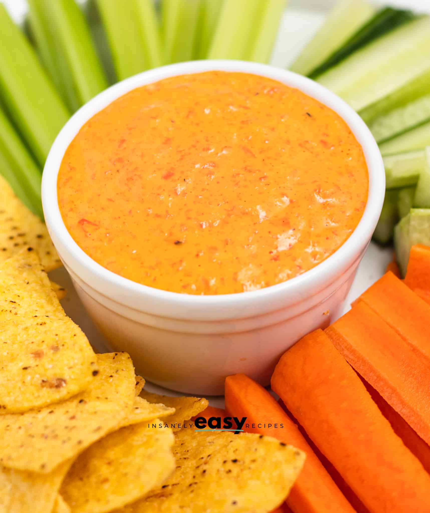 closeup view of a bowl of red pepper crema surrounded by tortilla chips, carrot sticks, and celery for dipping. 