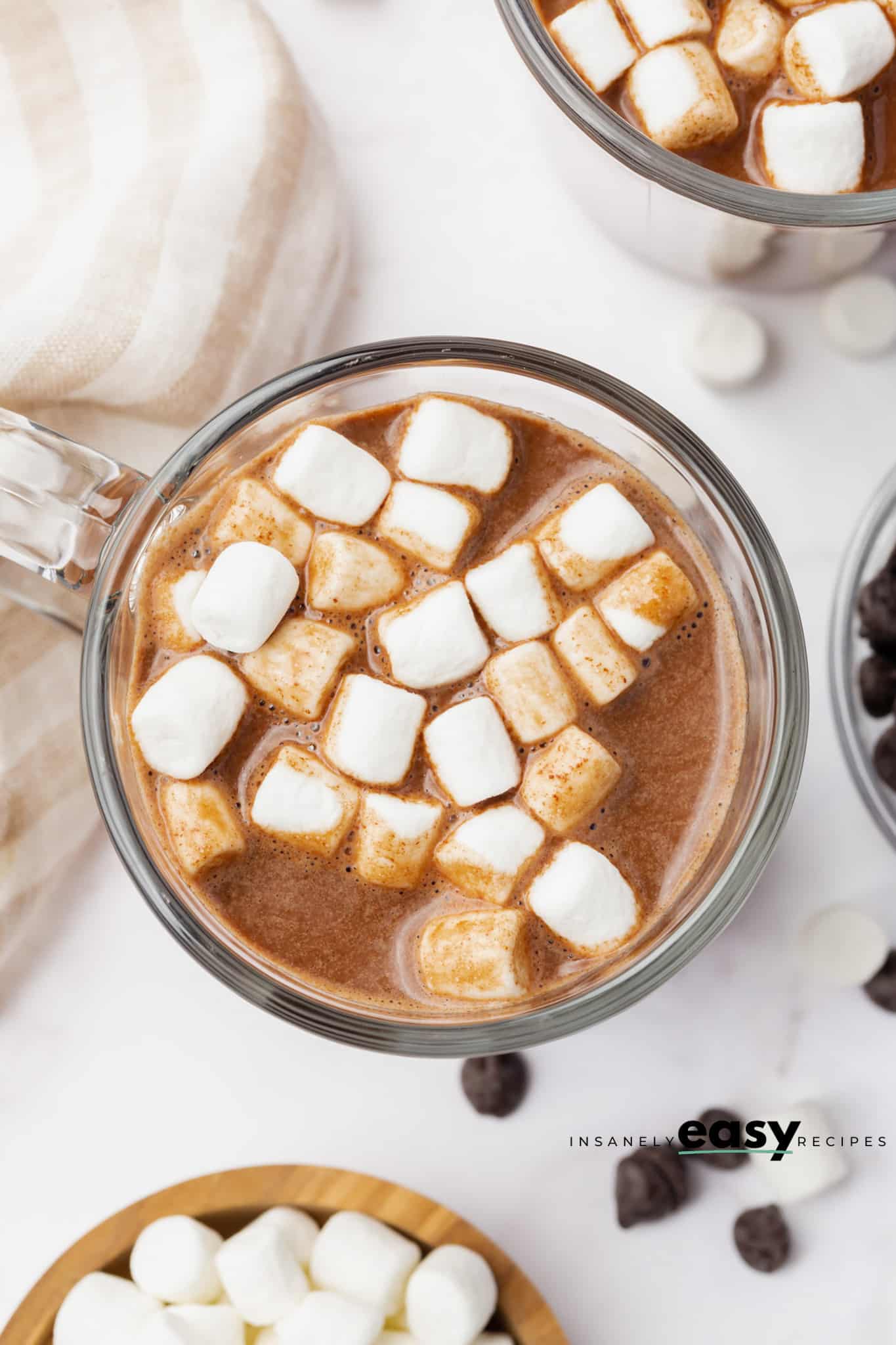 Top view photo of a glass mug filled with Vegan Hot Chocolate and mini marshmallows. There is a wooden bowl with marshmallows below the mug and another mug of hot chocolate above the center mug. 