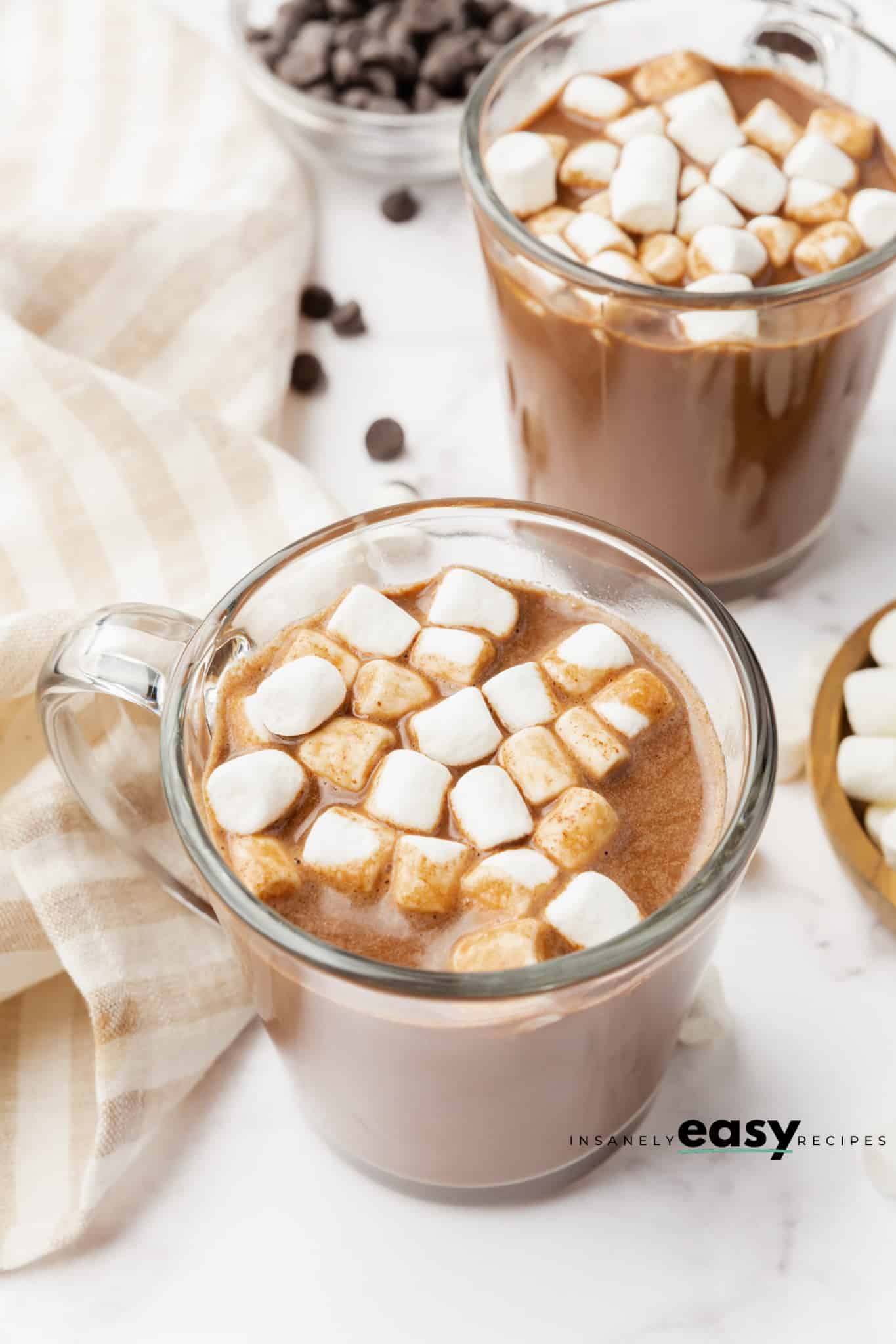 Photo of a glass mug willed with Vegan Hot Chocolate and mini marshmallows. There is a kitchen towel to the left of the mug, and another mug of hot chocolate behind the center mug. 