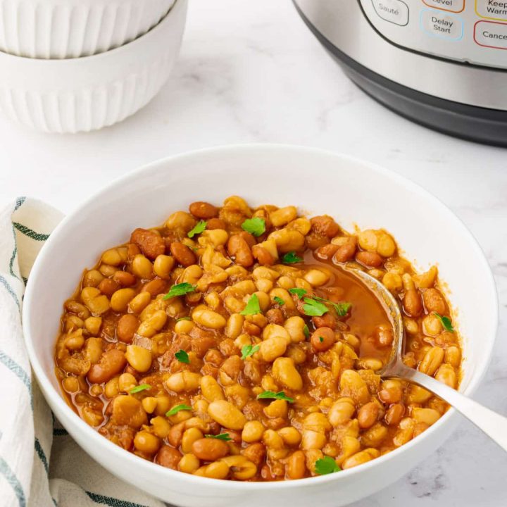 Photo of a white bowl with instant pot baked beans in the bowl, with a spoon in the bowl. It's garnished with freshly chopped parsley, and the instant pot is in the background.