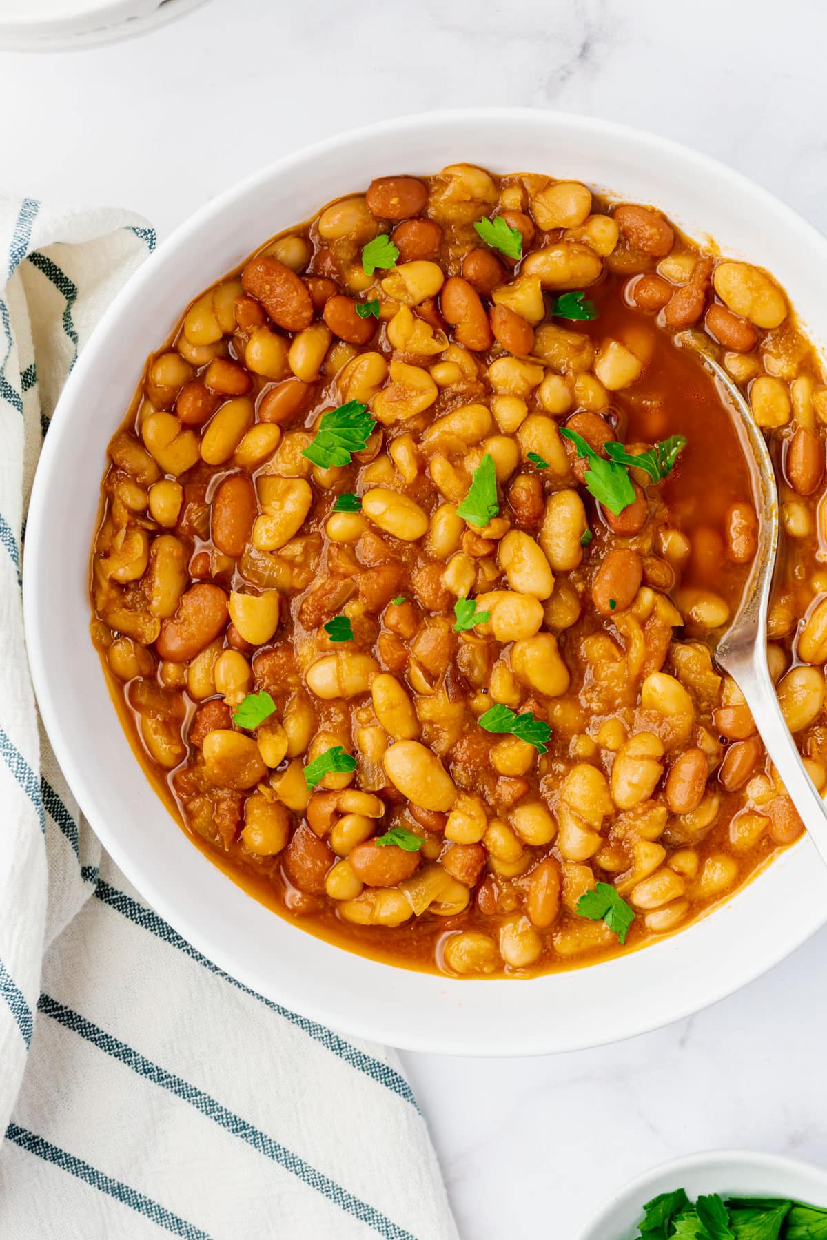 Top view photo of a white bowl filled with Instant Pot Baked Beans. There is a spoon in the bowl, and a kitchen towel by the bowl.