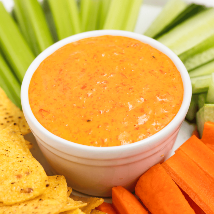 closeup view of a bowl of red pepper crema with tortilla chips and veggies on the side for dipping.