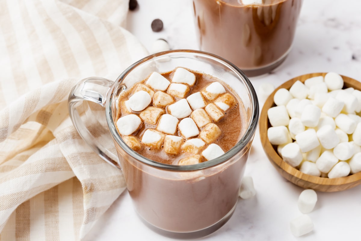 Photo of Vegan Hot Chocolate in a glass mug, with vegan marshmallows on top. There is a wood bowl with marshmallows to the right of the mug and a kitchen towel to the left of the mug.