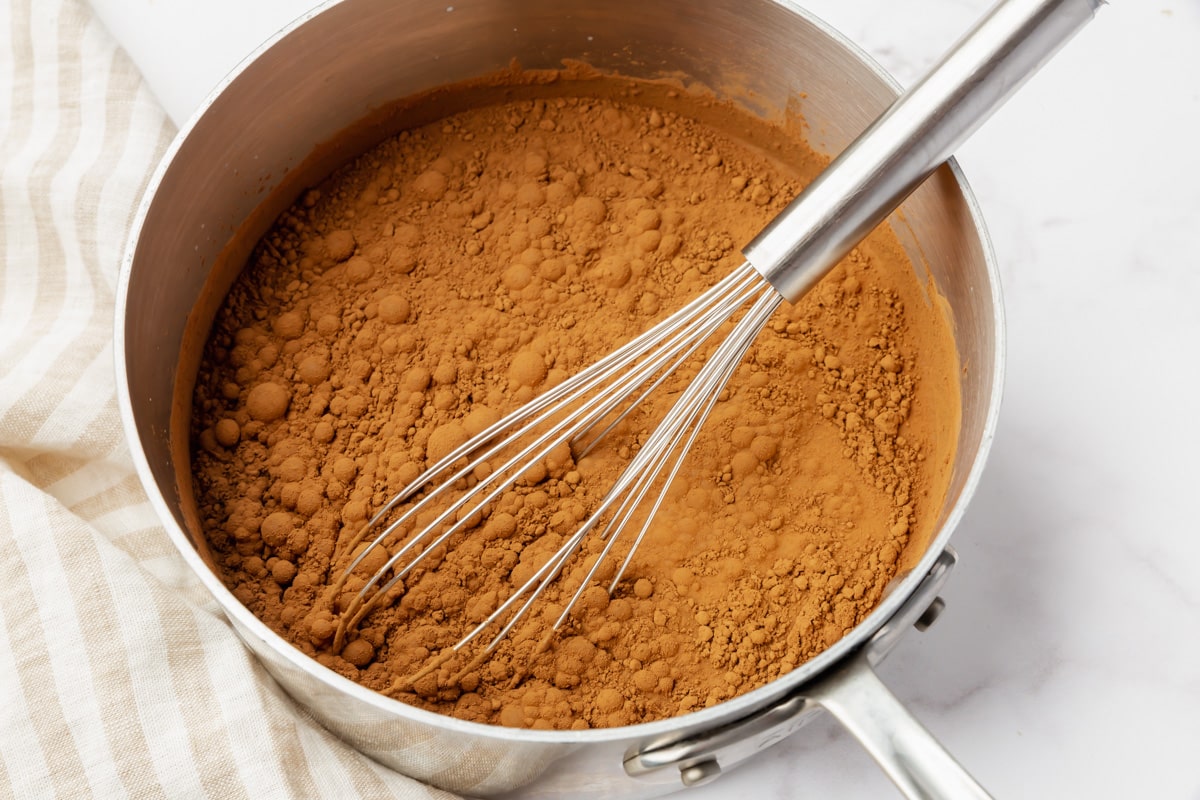 Top view photo of a saucepan with almond milk, vegan chocolate chips, cocoa powder, and granulated sugar, ready to mix with a wire whisk.