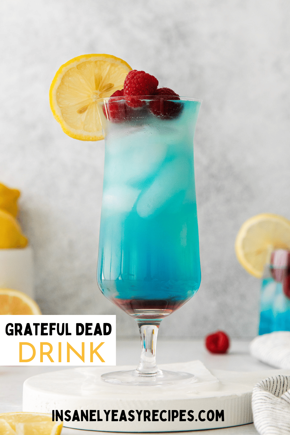 tall clear glass with red liquid at bottom, then blue liquid, with rapsberries and blue umbrella in drink. Surrounded by lemons and raspberries on the table with text overlay: grateful dead drink