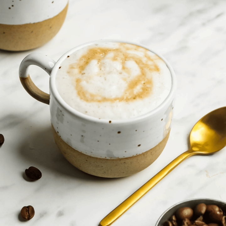 Closeup photo of two mugs filled with Oat milk Latte. There is a gold spoon next to the mug, a measuring cup filled with coffee beans next to the spoon.