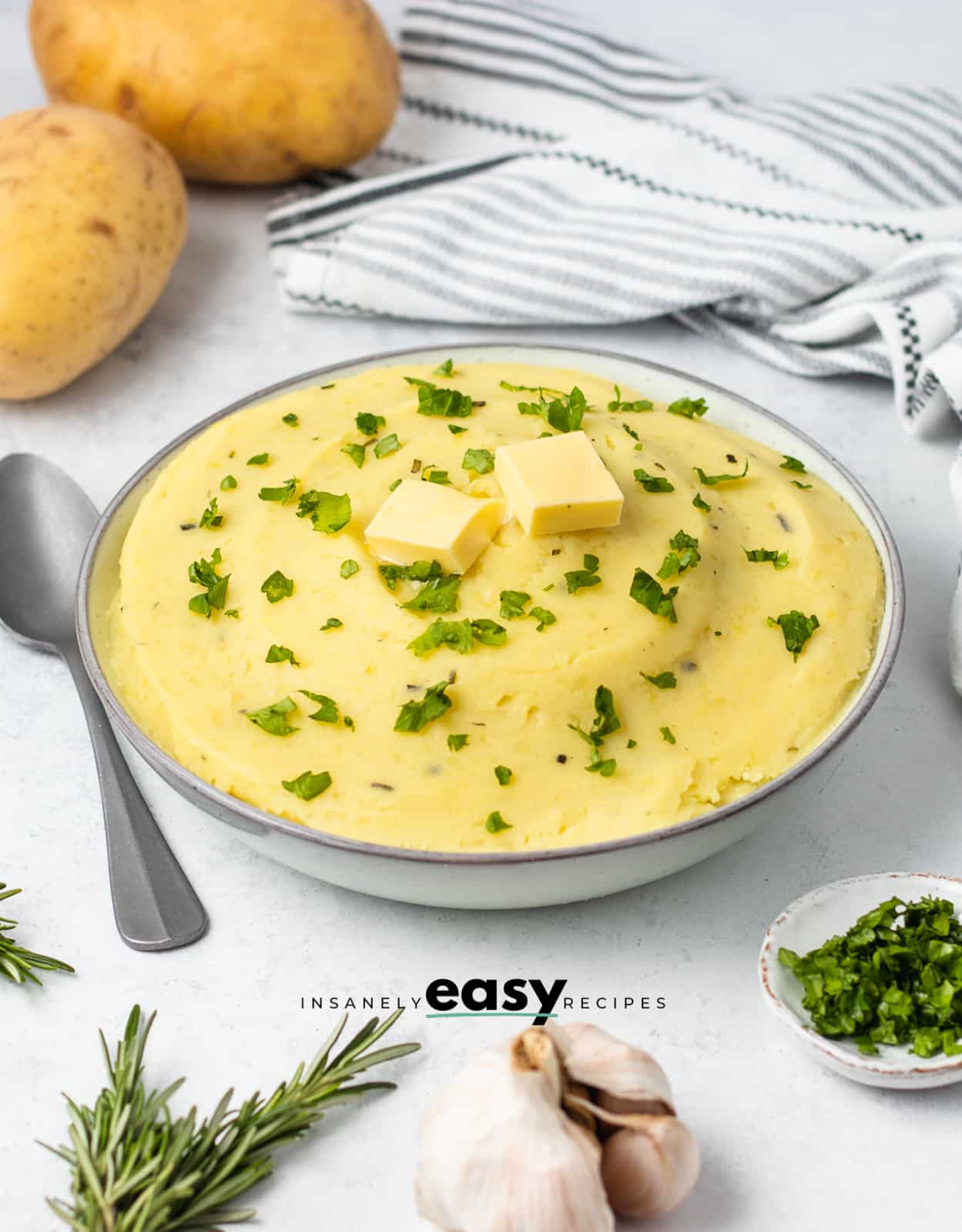 Photo of a large white bowl filled with Rosemary Mashed Potatoes, and finished with fresh parsley and two pats of butter. 
