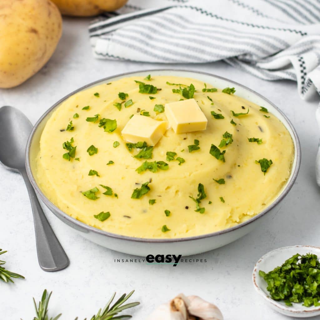 Photo of a large white bowl filled with Rosemary Mashed Potatoes, and finished with fresh parsley and two pats of butter.