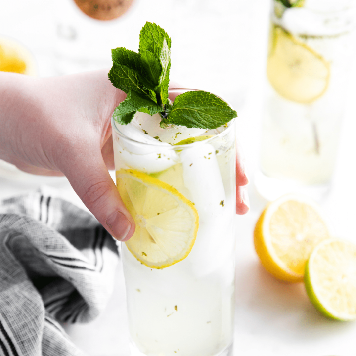 a hand picking up a bootlegger drink in a tall glass, garnished with lemon and mint.