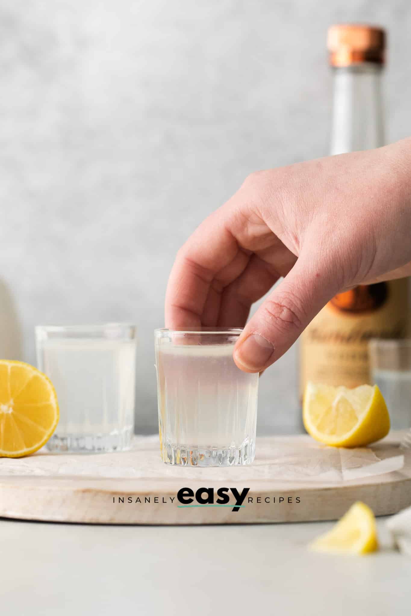 Photo of a hand grabbing a White Tea Shot. There are two shots on a wooden platter with lemon wedges around the shots. 