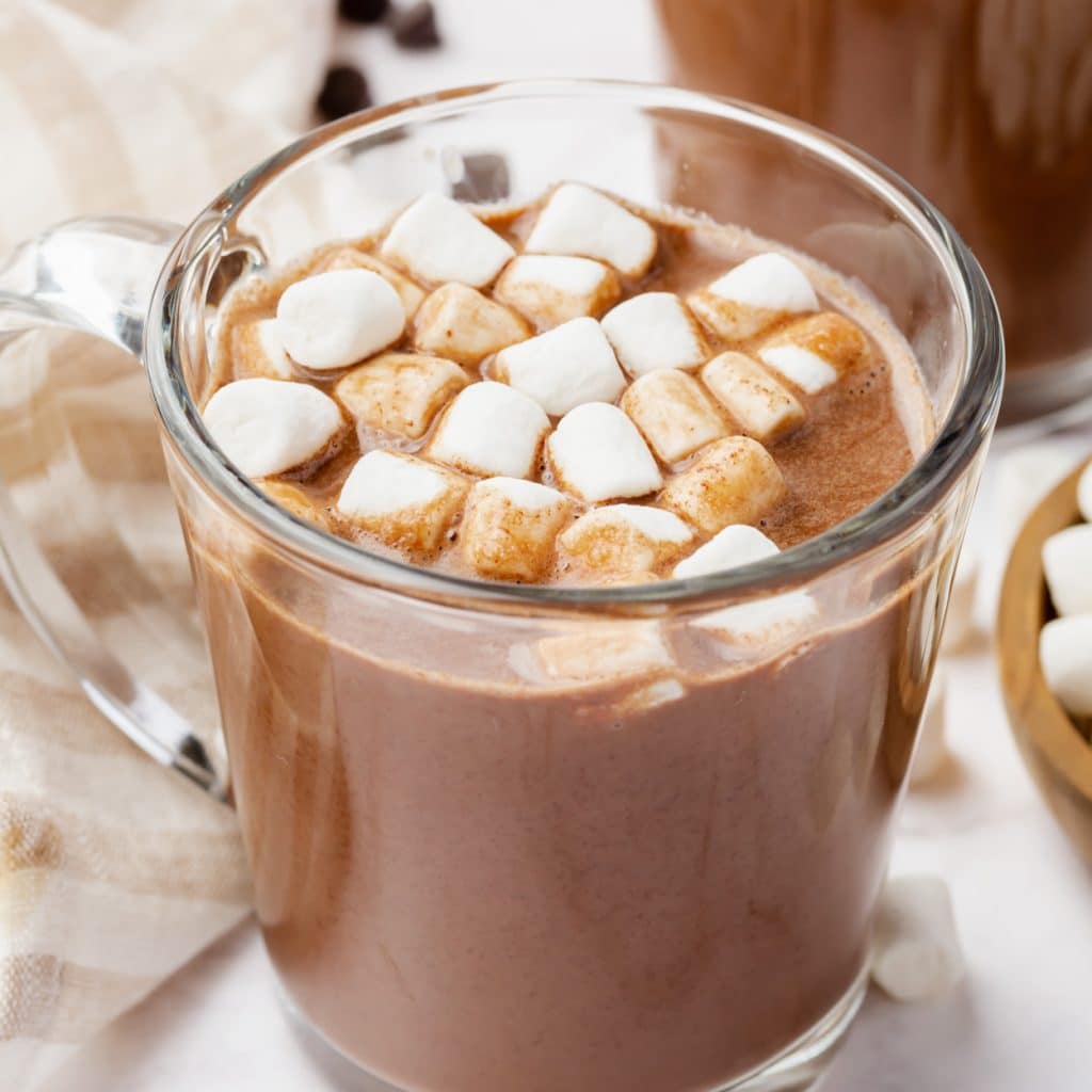 Photo of Vegan Hot Chocolate in a glass mug, with vegan marshmallows on top. There is a wood bowl with marshmallows to the right of the mug and a kitchen towel to the left of the mug.