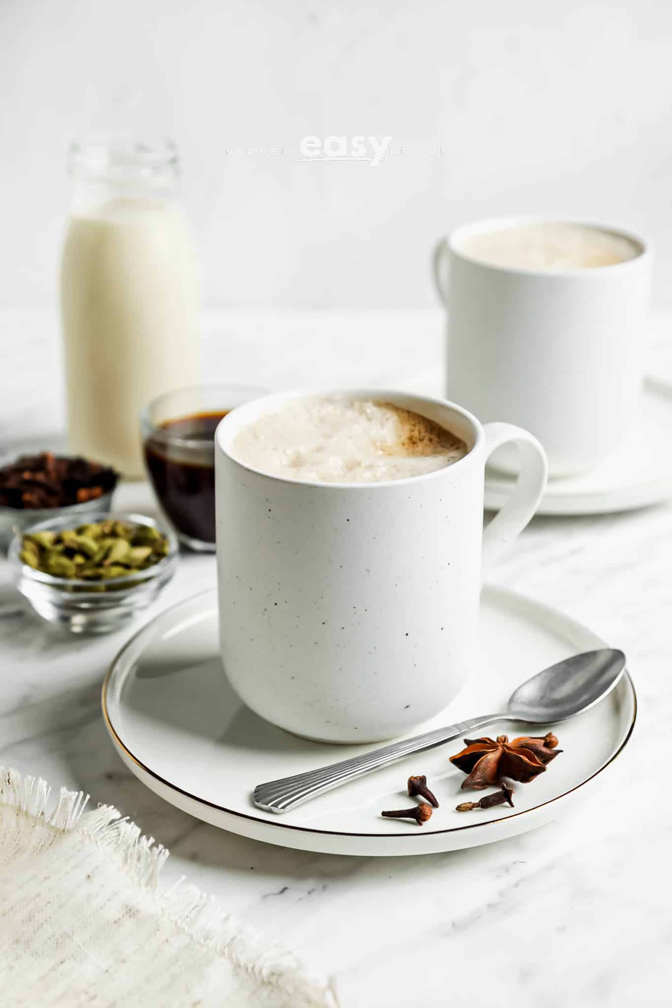 Photo of a white mug filled with a Dirty Chai Latte. There are small bowls with cardamom pods, star anise, and black tea around the mug.
