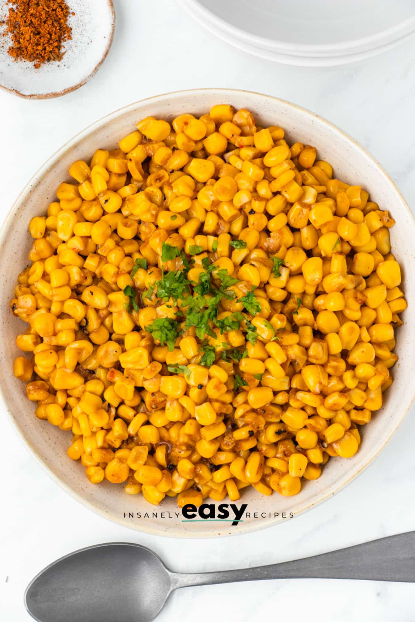 a large ceramic serving bowl filled with yellow corn with cajun seasonings and parsley garnish. There is a spoon under the bowl and a small plate of cajun seasoning above it. View is from overhead.