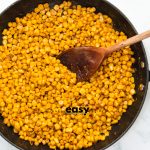 A skillet of yellow corn kernels and cajun seasonings, stirred with a wooden spoon.