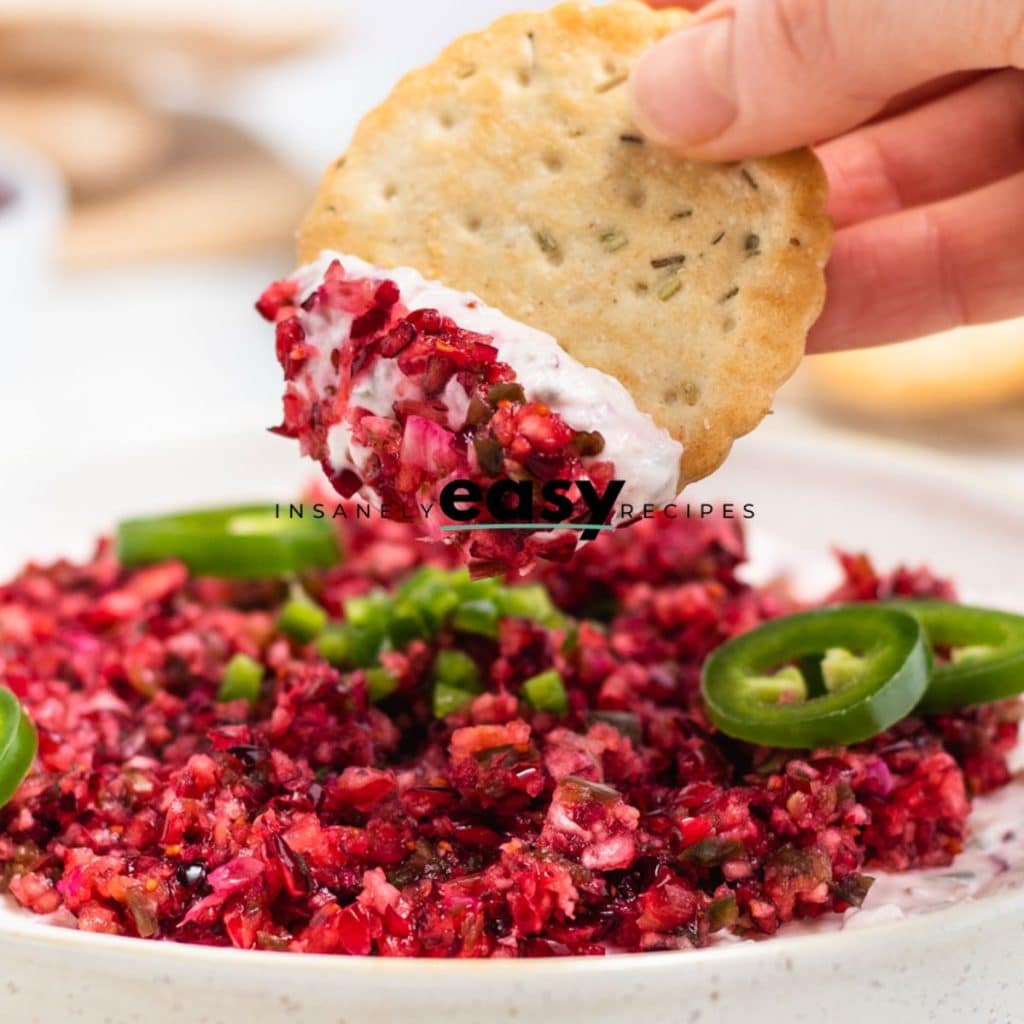 Closeup photo of a hand dipping a cracker into cranberry jalapeno dip in a white bowl.