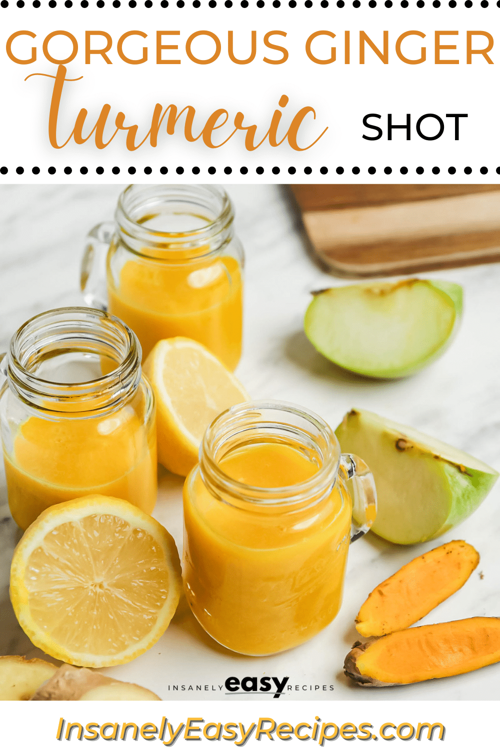 a small mason jar mug filled with orange ginger turmeric juice. Around the jar are pieces of apples, lemon, ginger, and turmeric root. text overlay at top of image says "gorgeous ginger turmeric shot"
