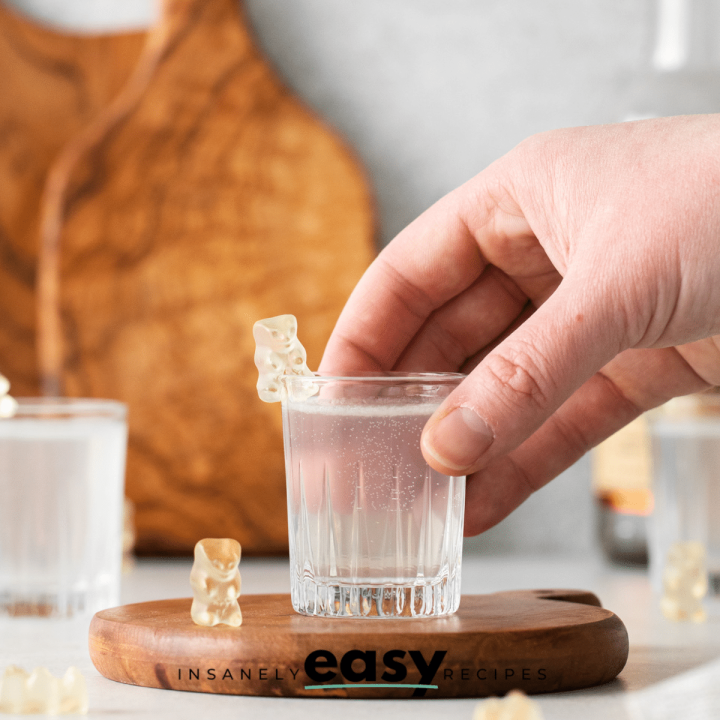 Photo of a hand grabbing a White Gummy Bear Shot. The shot glass is on a wooden coaster surrounded by white gummy bears.
