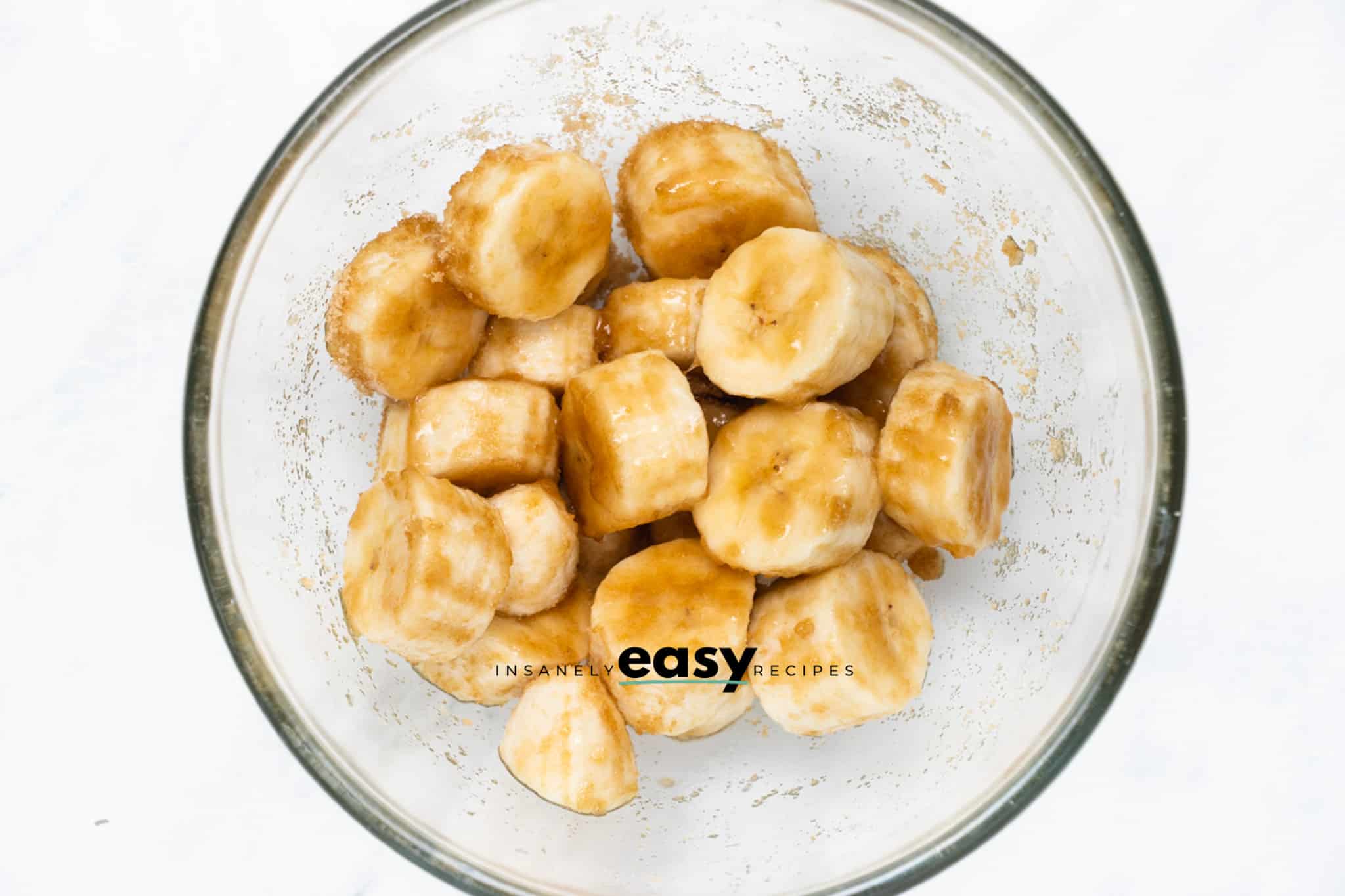 Top view photo of sliced bananas in a glass bowl, tossed in brown sugar.