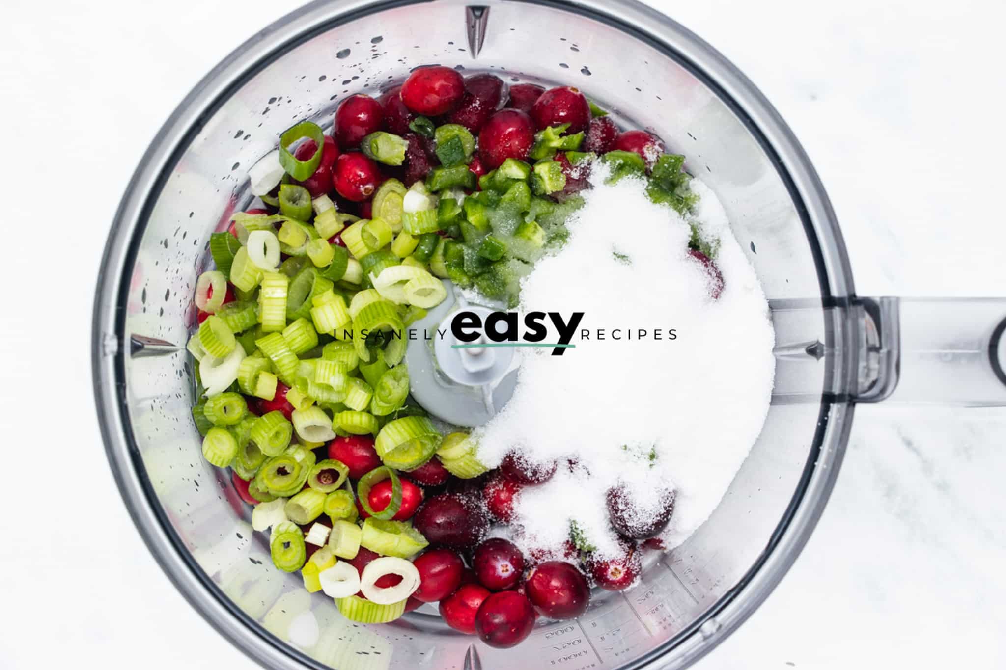 Top view photo of a food processor with cranberries, scallion, jalapeno, lime juice, sugar and salt, ready to chop until fine.