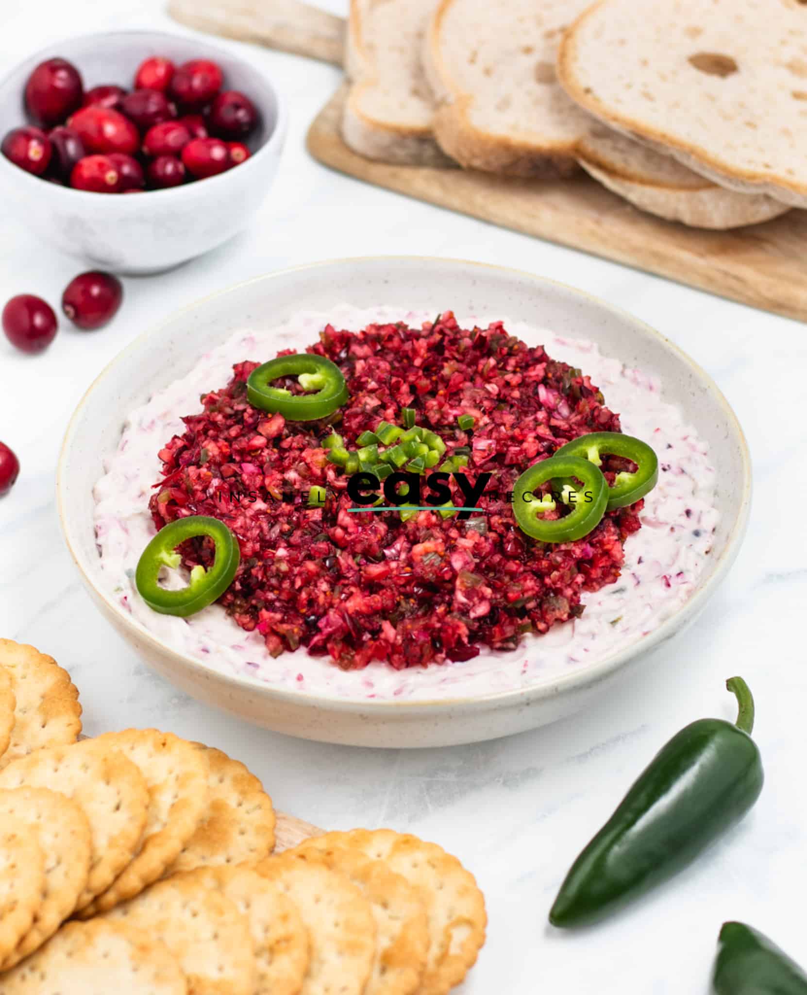 Photo of a white bowl filled with Cranberry Jalapeno Dip. The bowl is surrounded by a fresh jalapeno, cranberries, and crackers. 