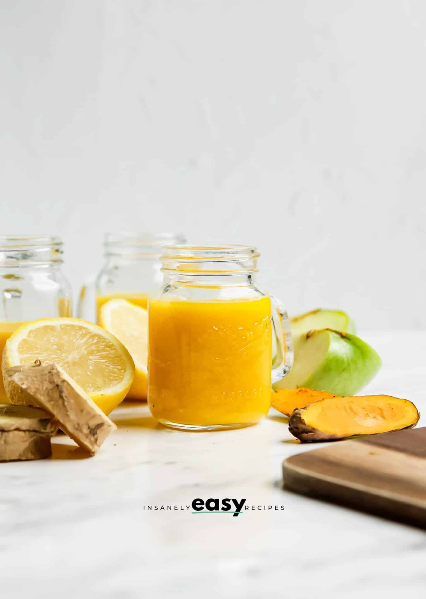 a small mason jar style shot glass with a handle, filled with blended ginger turmeric juice. In the background are apples, lemons, and turmeric roots. 