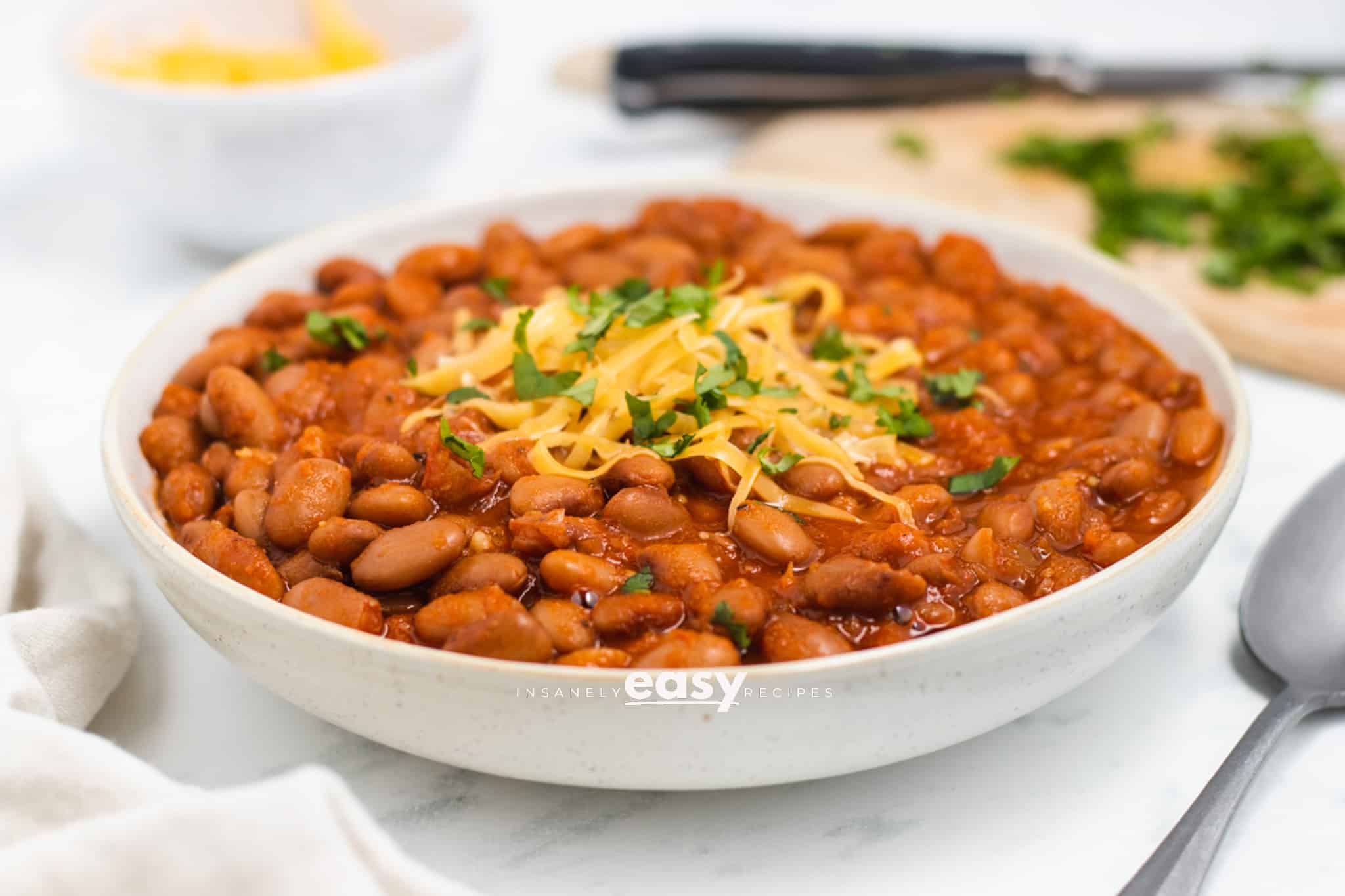 a bowl filled to the top with seasoned ranch style beans. They are topped with shredded cheese and herbs.