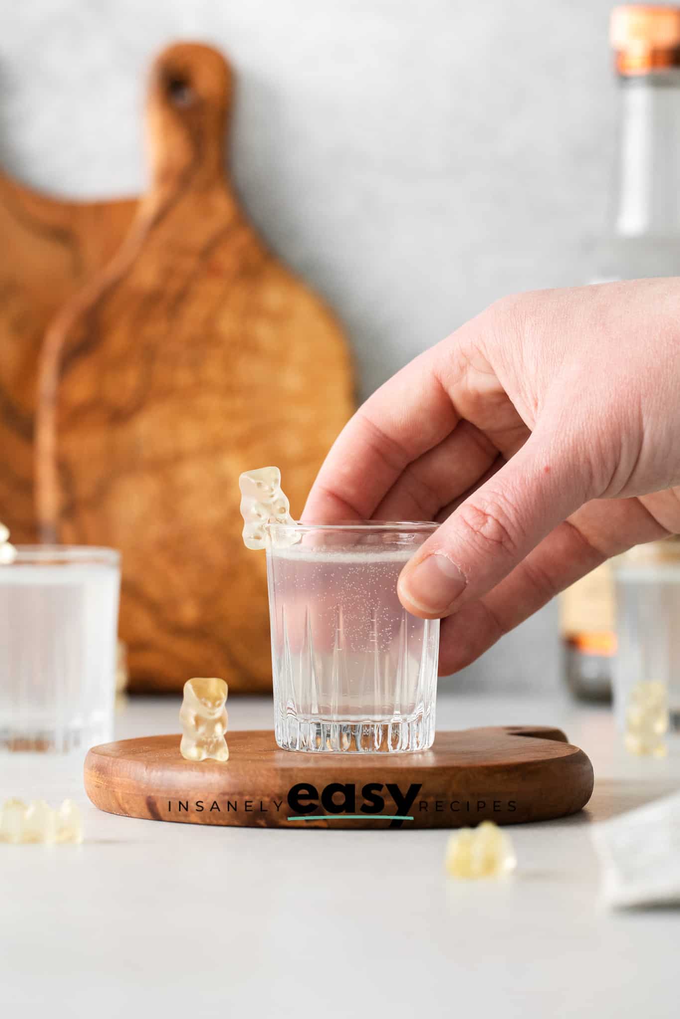 Photo of a hand grabbing a White Gummy Bear Shot. The shot glass is on a wooden coaster and surrounded by white gummy bears. 
