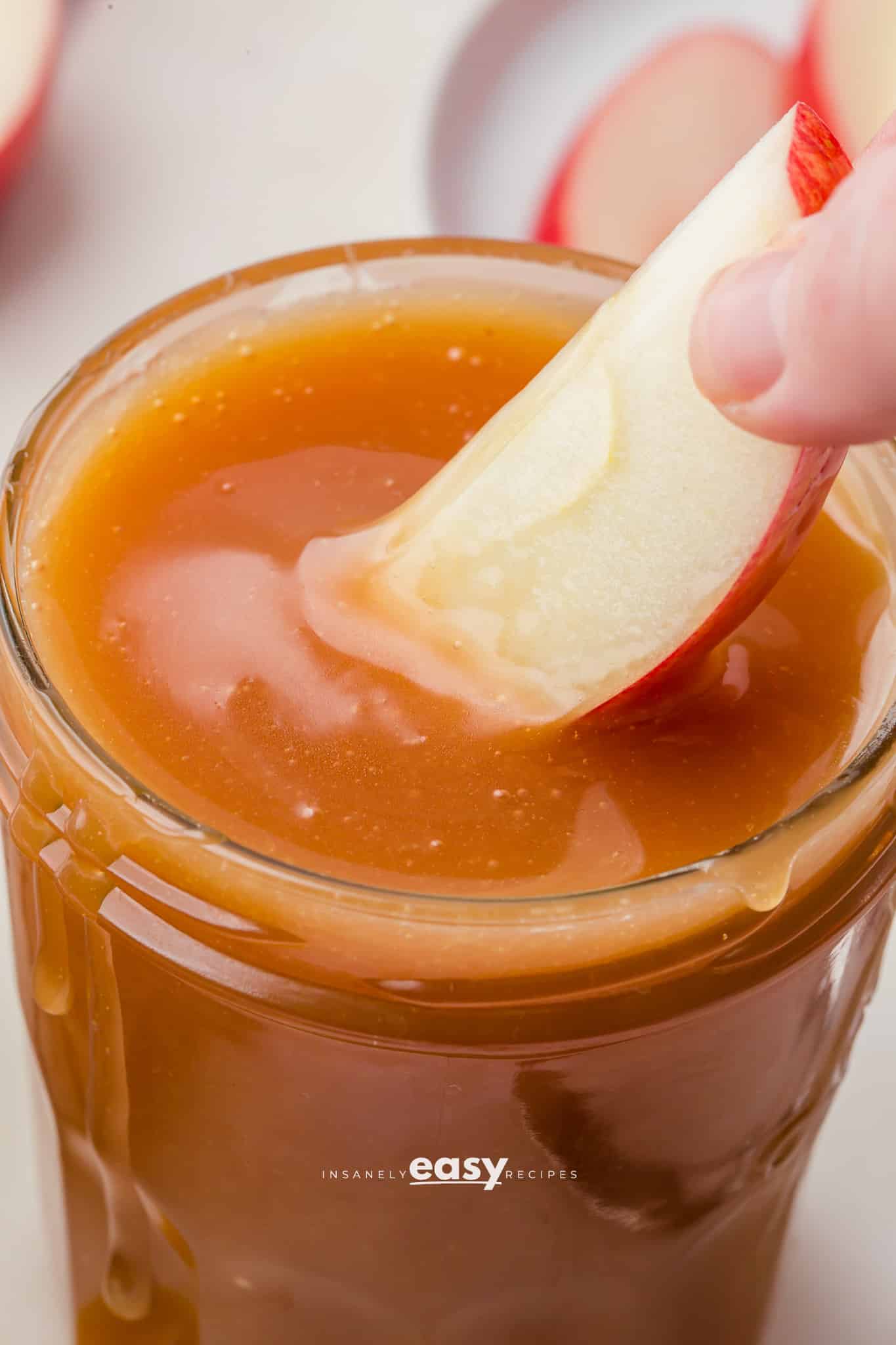 An apple slice dipping into a small glass jar filled with homemade microwave caramel sauce