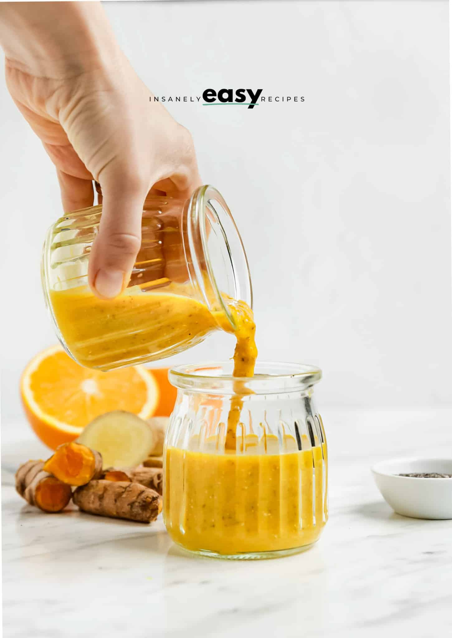 Photo of a hand pouring a wellness shot from a glass jar into another small glass jar. There is an orange cut in half in the background as well as fresh ginger and a bowl of chia seeds.