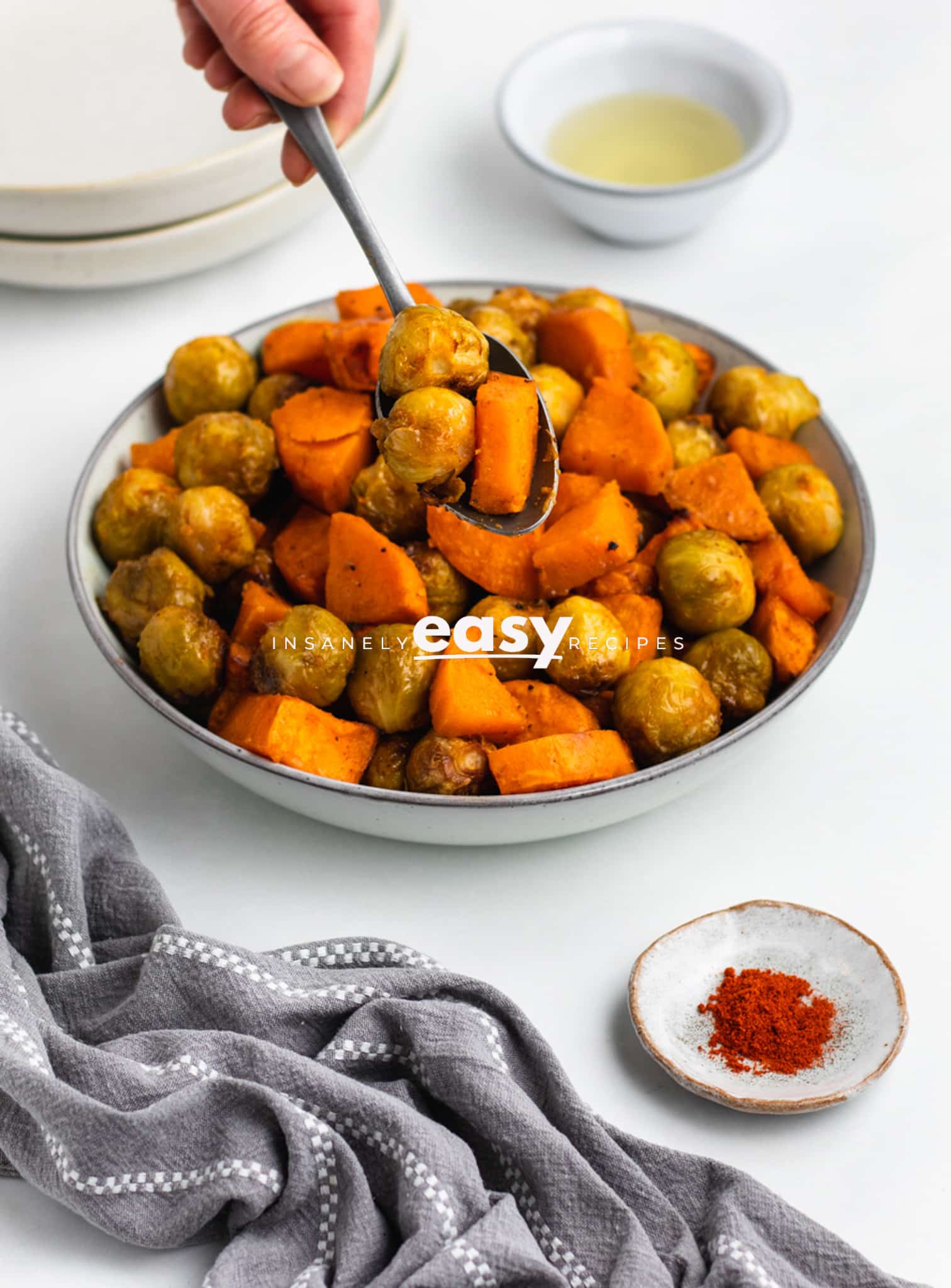 Photo of a hand holding a spoon, and scooping freshly roasted brussel sprouts and sweet potatoes out of a white bowl. 
