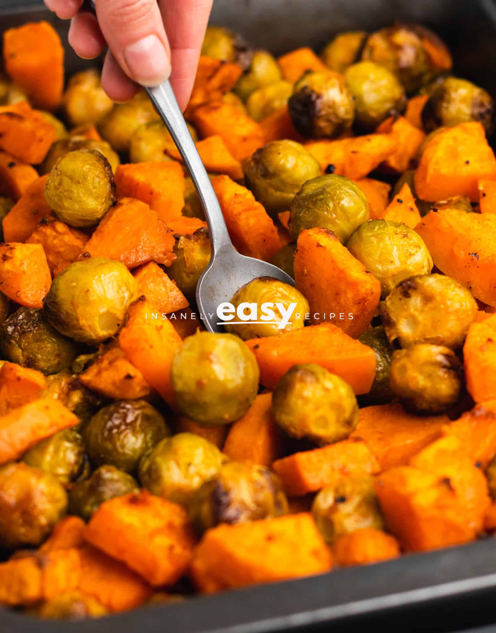 Closeup photo of a hand scooping up roasted brussel sprouts and sweet potatoes from a sheet pan. 