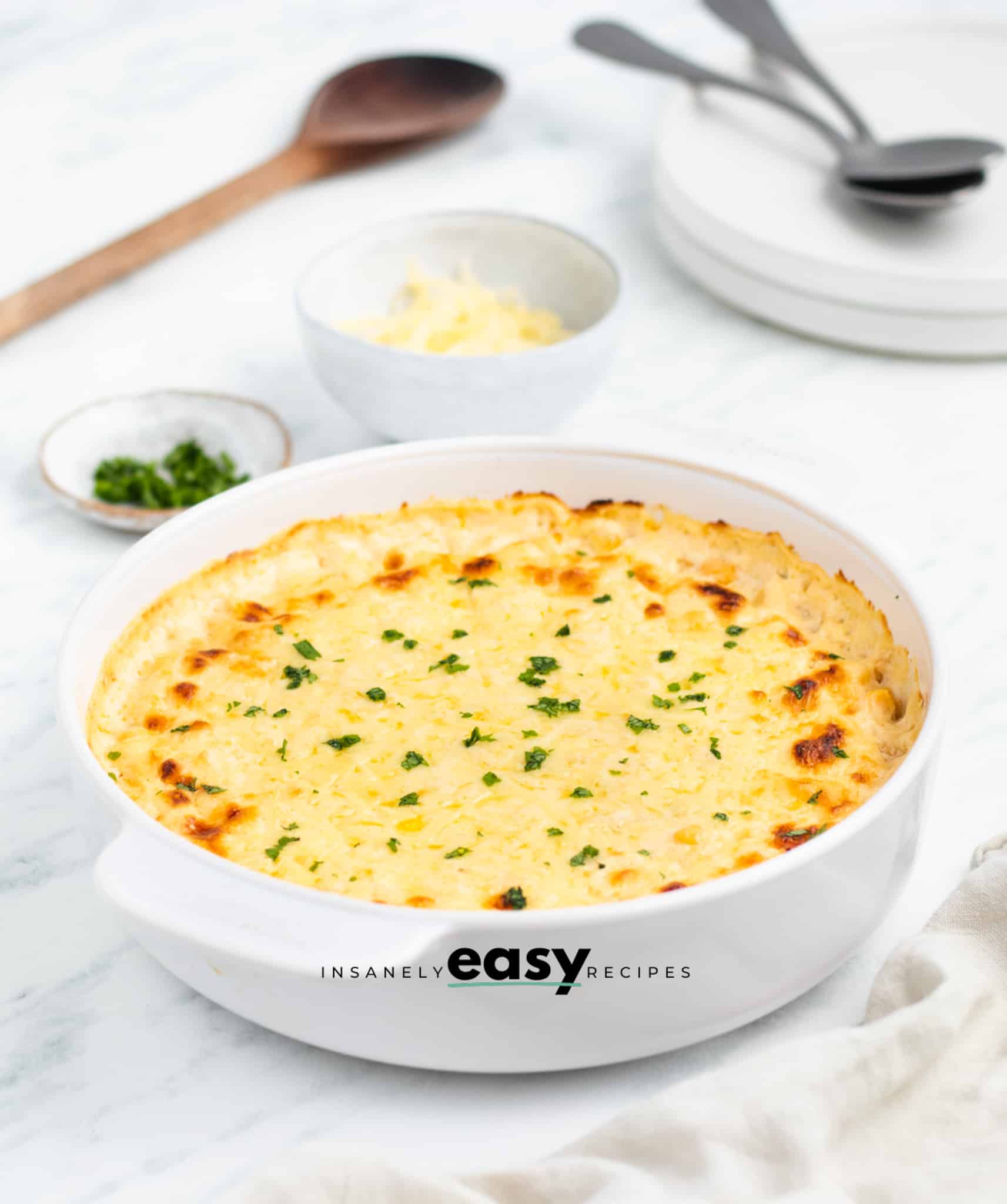 Photo of corn casserole with cream cheese in a white casserole dish, ready to serve. There is a wooden spoon and silver spoons in the background. 