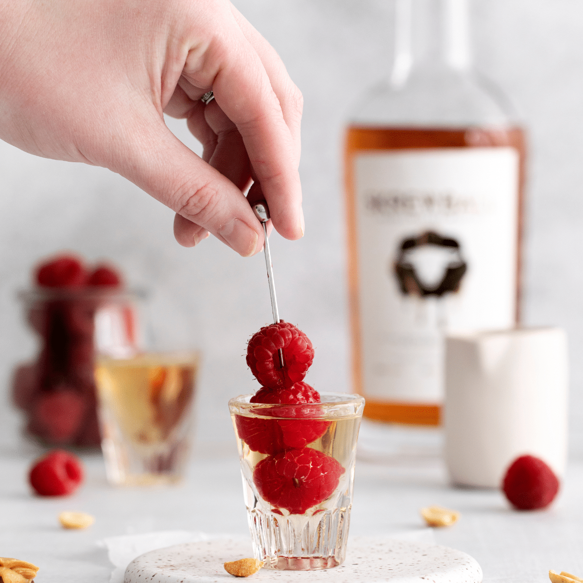 Photo of a PB and Jelly Shot in a shot glass, with a hand place 3 raspberries on a toothpick in the glass. There is peanut butter liquor in the background.