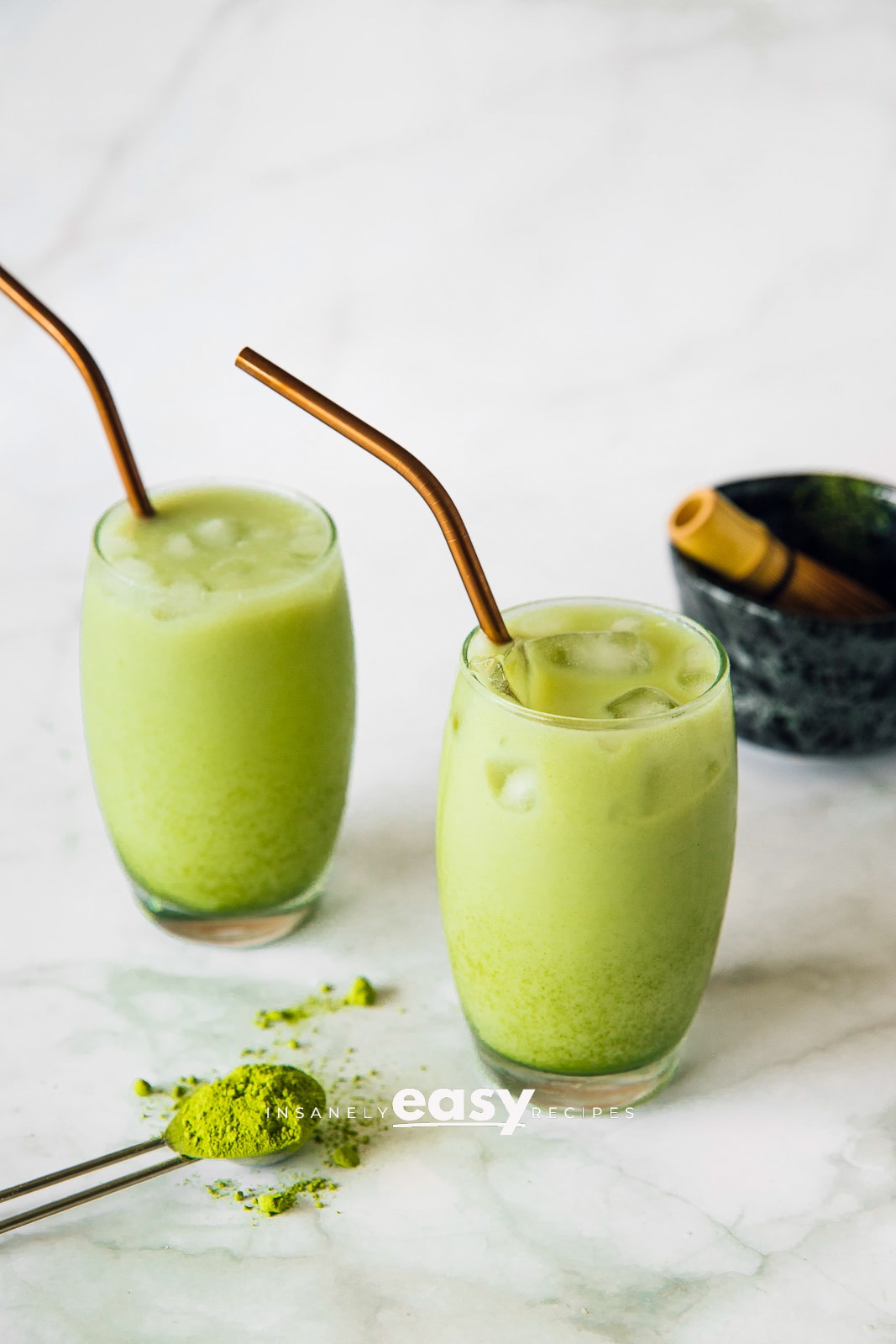 Photo of two glasses filled with matcha latte. There is a teaspoon with dried matcha powder in the foreground and a small bowl with a matcha mixer in the background. 