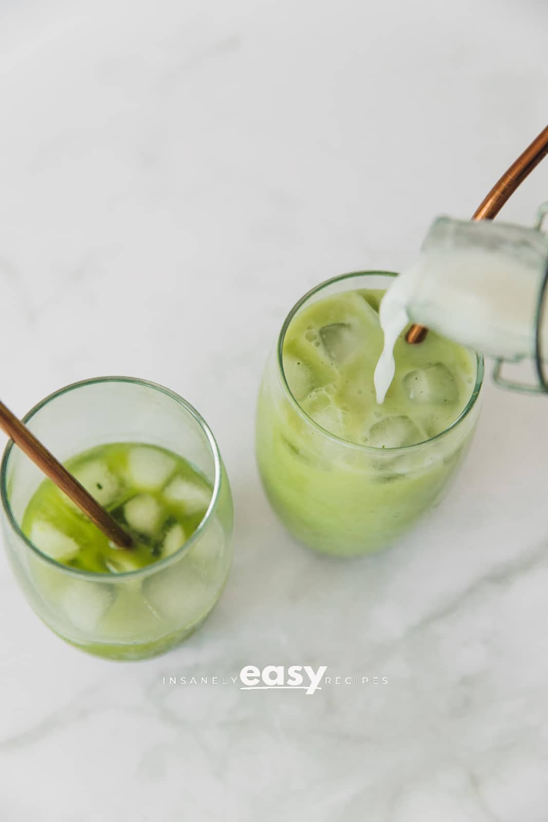 Top view photo of coconut milk being poured into a glass with ice and matcha. The glasses have metal straws. 