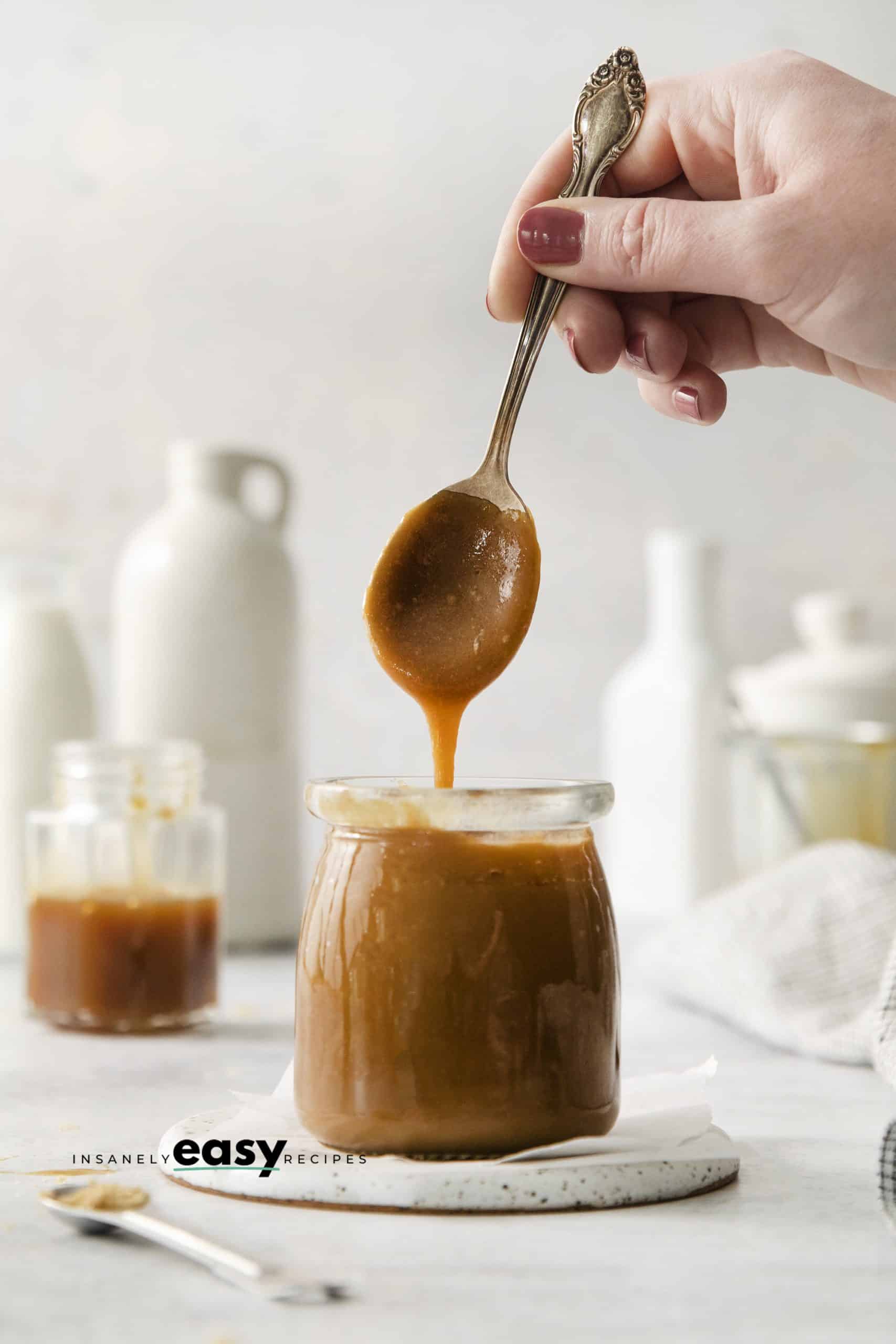 a hand holding a spoon of caramel sauce over a jar of the same.